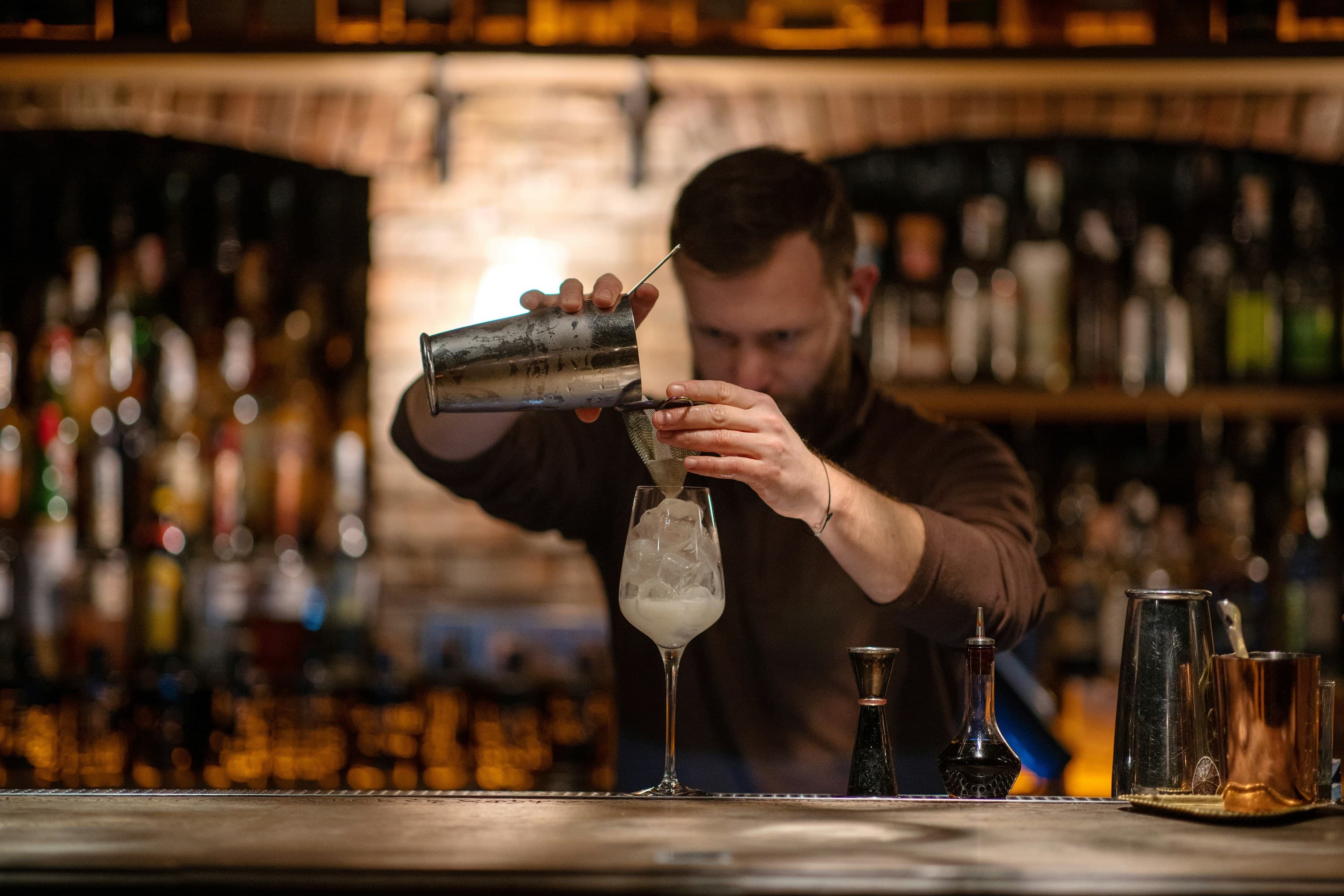 a bartender straining a shaken drink into a stemmed glass against warm exposed brick, bottles in amber blur behind