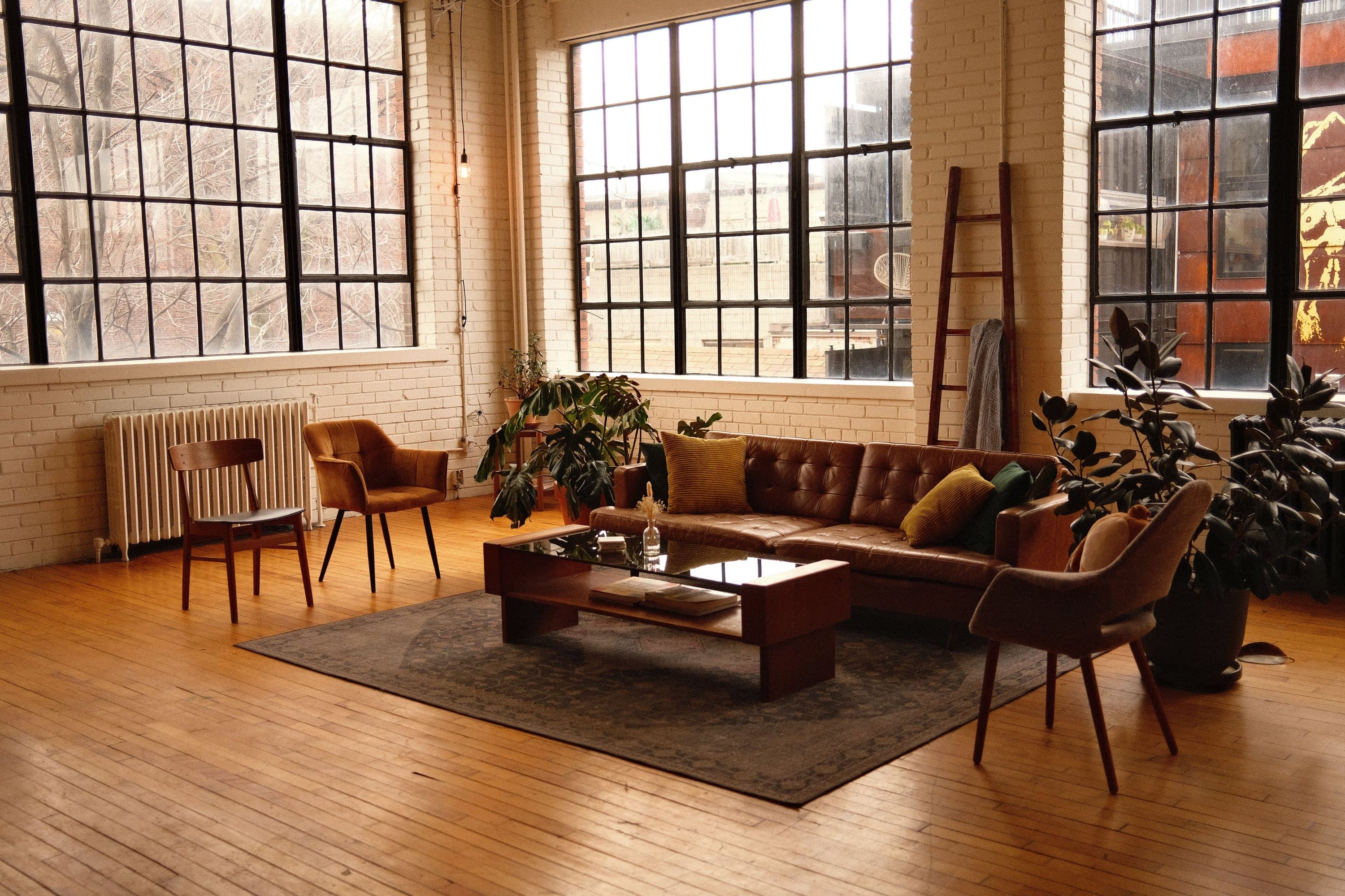 a warehouse loft in late light, steel windows over painted brick, a leather sofa and chairs on a worn rug