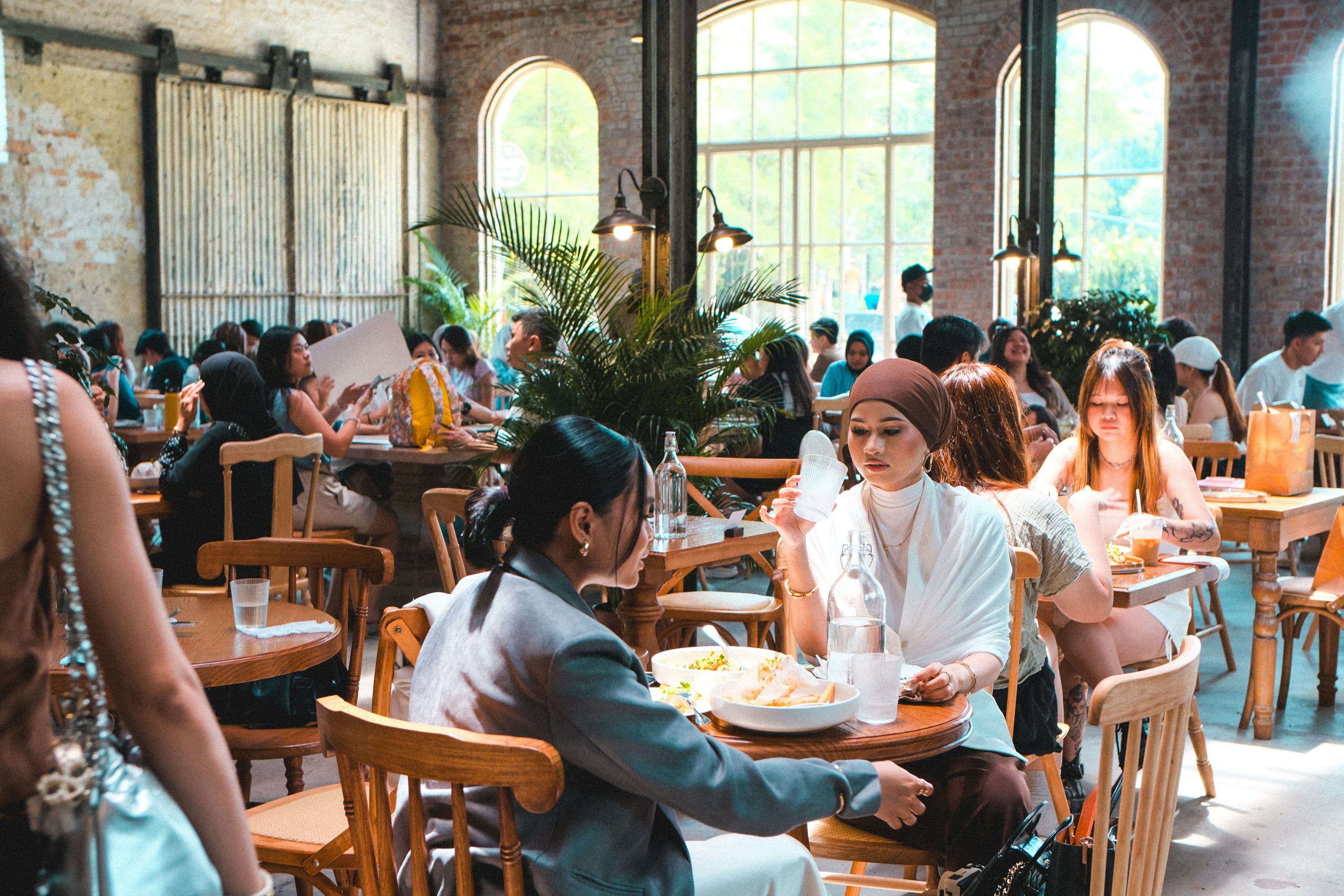 a packed brick warehouse hall under tall arched steel windows and palms, people at small wooden tables talking and eating