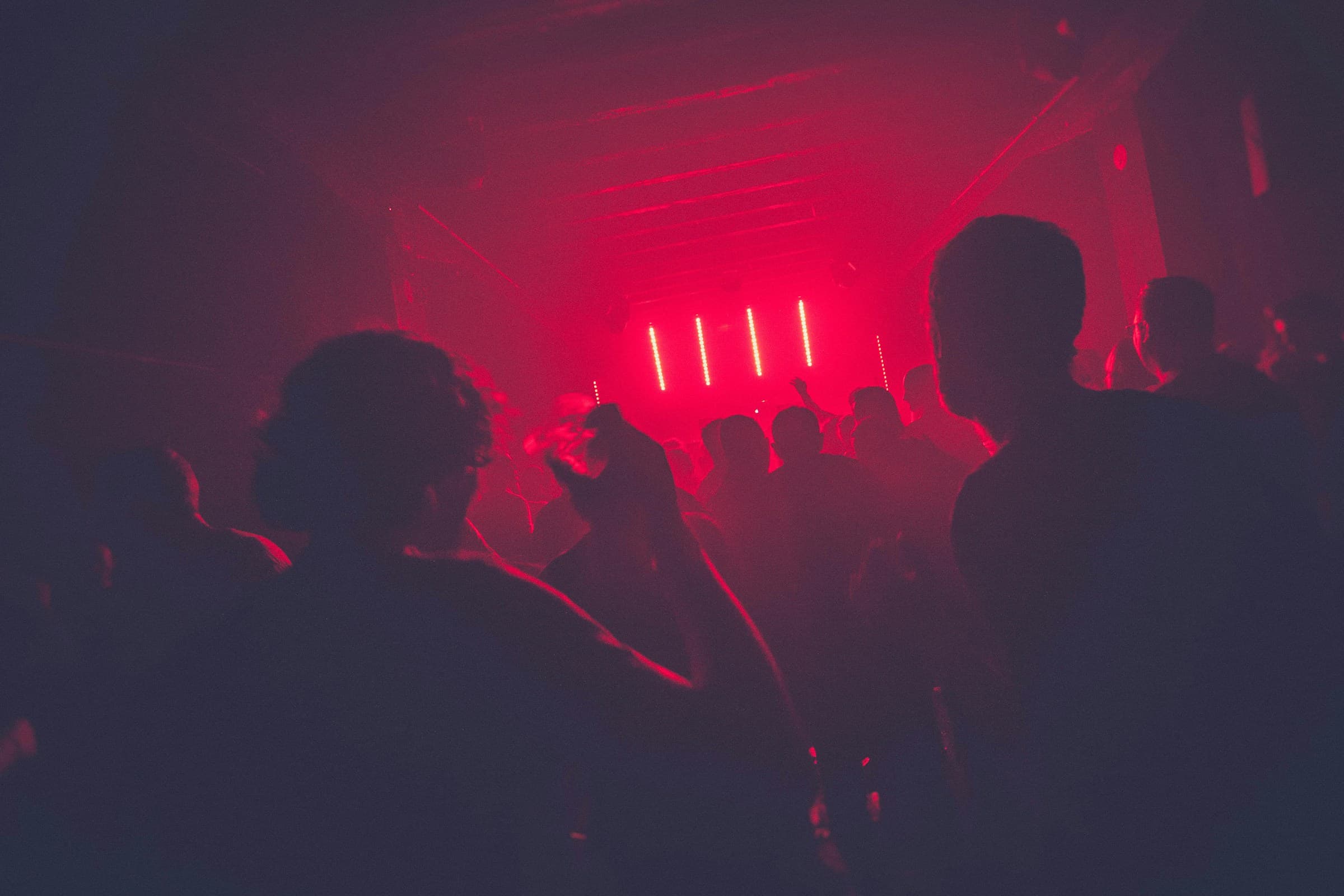 a packed low room late, shoulders and raised hands in deep red light, a line of lamps at the far end