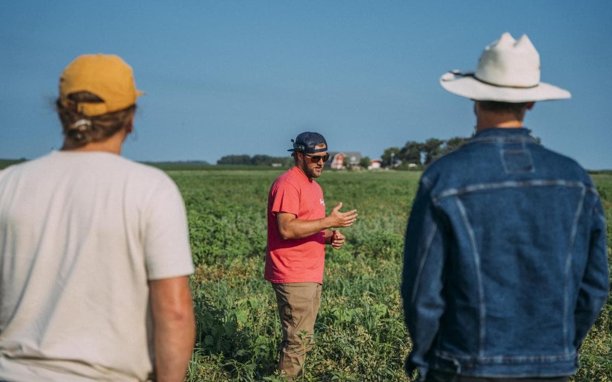 a farmer in a red shirt mid-sentence in a green field, two listeners in the foreground, one in a white cowboy hat