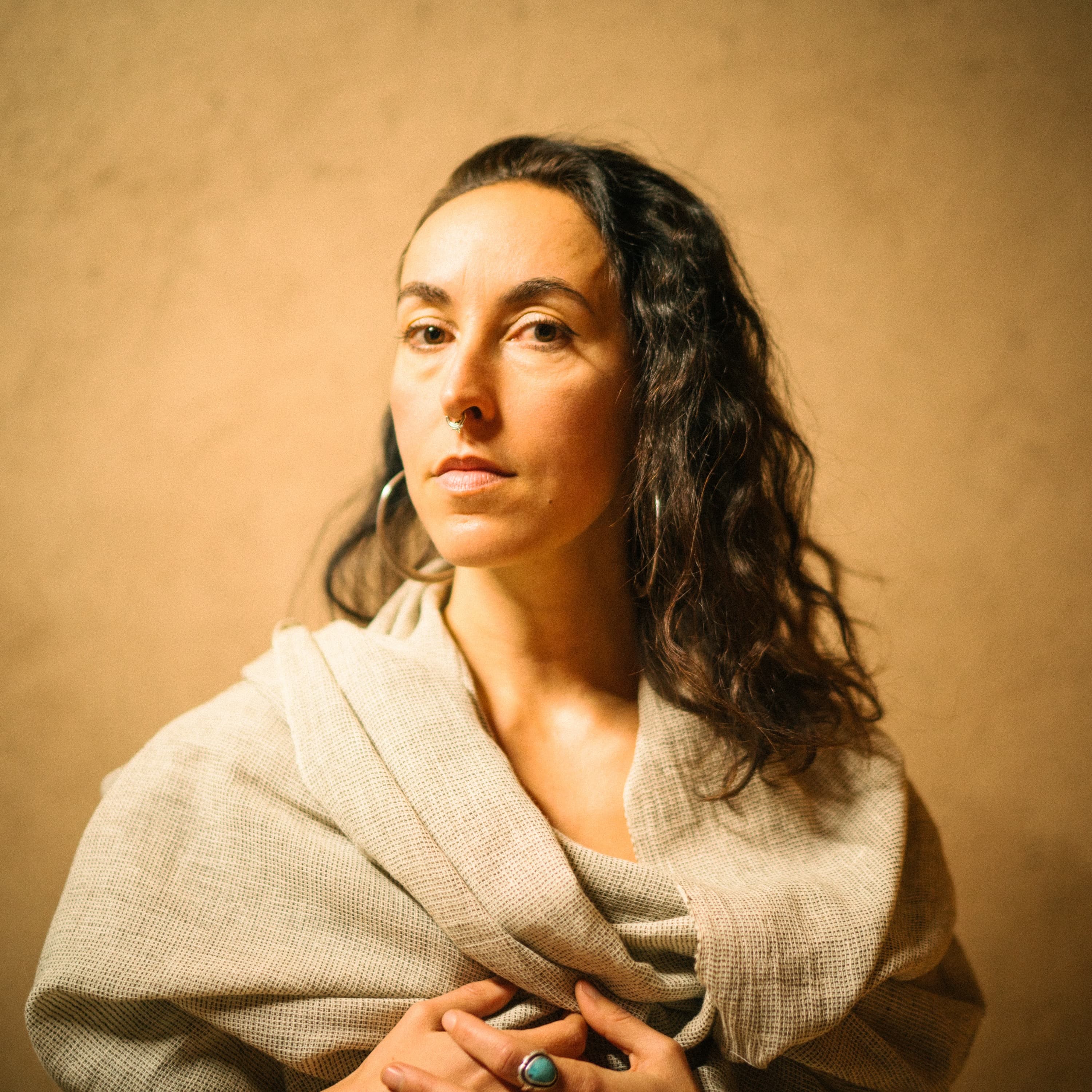 rising appalachia, a singer in a linen wrap against a warm plaster wall, hands folded at her chest